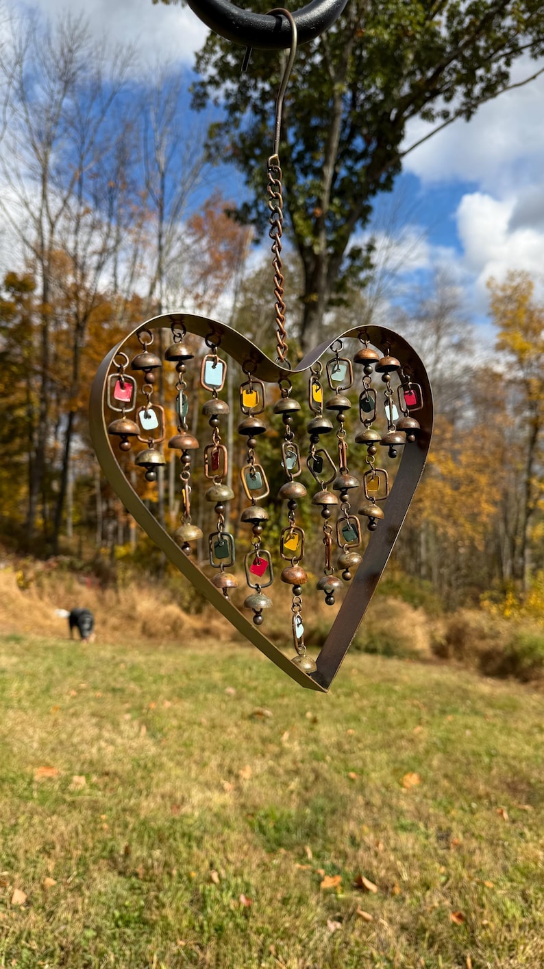 Handcrafted heart wind chime featuring colorful hanging dangles and decorative metal bells with a beautiful flamed finish. Metal wind chimes outdoor designed for garden decor, patio accents, porch decoration, balconies, fences, and backyard landscapes. Heart-shaped hanging garden ornament creates gentle, relaxing sounds while adding artistic charm to outdoor spaces. Perfect farmhouse decor, metal garden art, yard art, housewarming gift, romantic garden decor, and year-round outdoor decoration.