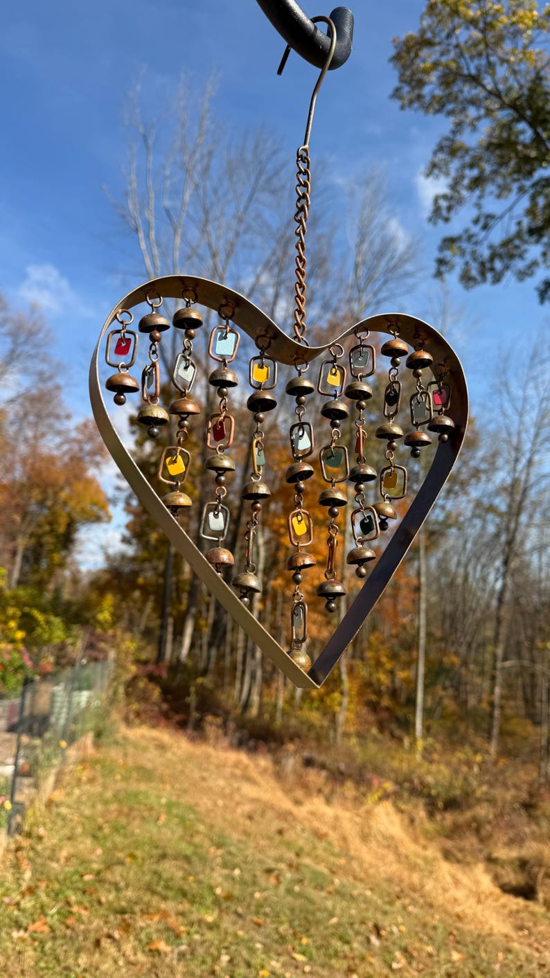 Handcrafted heart wind chime featuring colorful hanging dangles and decorative metal bells with a beautiful flamed finish. Metal wind chimes outdoor designed for garden decor, patio accents, porch decoration, balconies, fences, and backyard landscapes. Heart-shaped hanging garden ornament creates gentle, relaxing sounds while adding artistic charm to outdoor spaces. Perfect farmhouse decor, metal garden art, yard art, housewarming gift, romantic garden decor, and year-round outdoor decoration.
