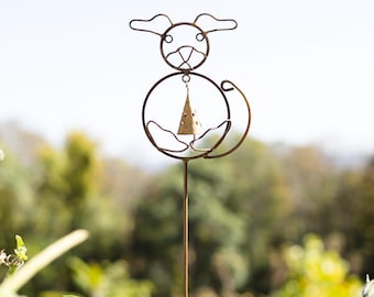 May include: A whimsical metal garden stake featuring a dog design. The dog's body is a circle with a bell hanging inside. The stake is made of a bronze-colored metal and is placed in a garden setting.