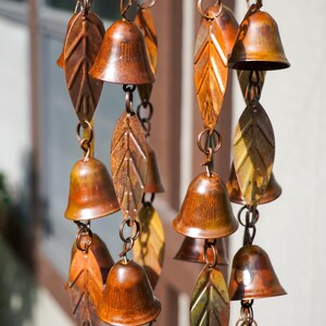 Puede incluir: Un carill&oacute;n de viento con m&uacute;ltiples campanas de metal en tono cobre y adornos en forma de hoja suspendidos de un marco circular. Los componentes est&aacute;n conectados por peque&ntilde;os anillos y cadenas de metal, creando una disposici&oacute;n vertical con acabado envejecido.