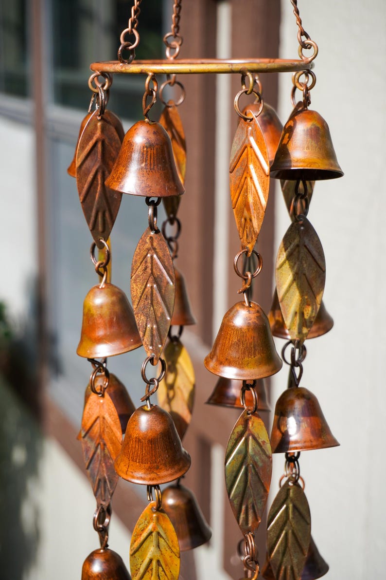 Puede incluir: Un carill&oacute;n de viento con m&uacute;ltiples campanas de metal en tono cobre y adornos en forma de hoja suspendidos de un marco circular. Los componentes est&aacute;n conectados por peque&ntilde;os anillos y cadenas de metal, creando una disposici&oacute;n vertical con acabado envejecido.