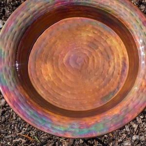 Hammered copper round birdbath with iridescent finish on curved metal stand.