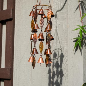Puede incluir: Un carill&oacute;n de viento colgante con m&uacute;ltiples campanas de metal en tono cobre y adornos en forma de hoja suspendidos de un marco circular mediante cadenas finas. El dise&ntilde;o incorpora varios tonos de marr&oacute;n met&aacute;lico y bronce en capas.