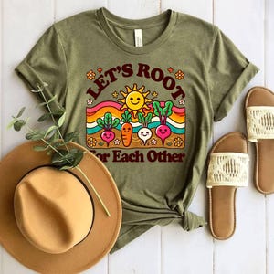 May include: Green short-sleeved t-shirt with a retro-style graphic of a sun, rainbow, and vegetables. The text on the shirt reads "Let's Root For Each Other".