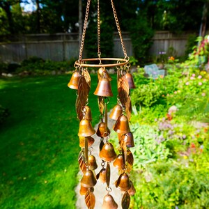 Puede incluir: Un carill&oacute;n de viento colgante con numerosas campanas de metal de color cobre y adornos en forma de hoja suspendidos de un marco circular mediante cadenas finas. La estructura se compone de varios niveles de campanas y hojas dispuestas.