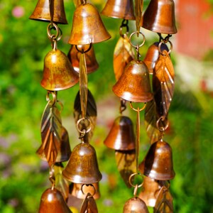 Puede incluir: Un carill&oacute;n de viento con m&uacute;ltiples campanas de metal de color cobre y colgantes en forma de hoja suspendidos de anillos circulares. Los componentes est&aacute;n dispuestos en hilos verticales, mostrando un acabado met&aacute;lico en tonos bronce y marr&oacute;n.