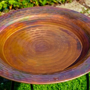 Hammered copper round birdbath with iridescent finish on curved metal stand.