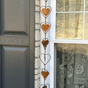 May include: A vertical hanging chain decoration featuring a series of connected metal hearts. Each heart is crafted from a dark, weathered copper-toned material and is framed by a thin, circular wire outline. The hearts are linked together by small metal rings.