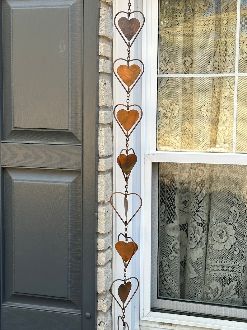 May include: A vertical hanging chain decoration featuring a series of connected metal hearts. Each heart is crafted from a dark, weathered copper-toned material and is framed by a thin, circular wire outline. The hearts are linked together by small metal rings.