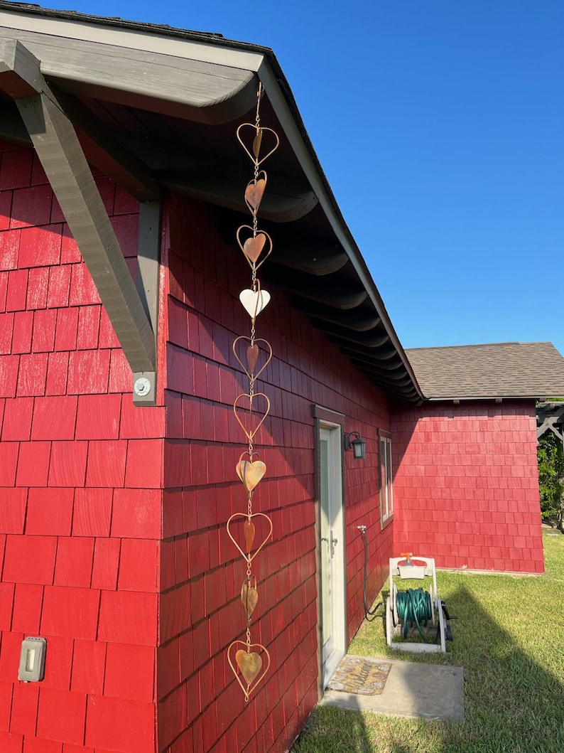 May include: A vertical rain chain hanging from the roof edge, constructed from a series of interconnected copper-colored metal hearts. The chain features alternating open heart frames and solid heart-shaped discs linked together in a long, linear design.