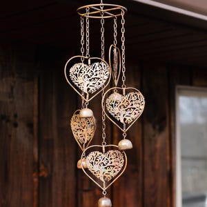 May include: A decorative wind chime with a bronze finish. The chime features heart-shaped and leaf-shaped cutouts with tree designs, suspended by chains. Small bells hang below each shape. The wind chime is hanging outdoors.