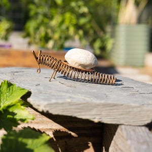 May include: A whimsical, handmade metal sculpture of a centipede with a coiled body and legs. The sculpture holds a smooth, light-colored stone on its back. The sculpture is placed on a gray stone surface, with a blurred green background.
