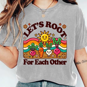 May include: A gray t-shirt with a retro-style graphic of a rainbow, sun, and cartoon vegetables. The text on the shirt reads "Let's Root For Each Other".