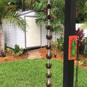 May include: A white shed with a brown door in a garden with a green lawn. A brown metal rain chain hangs from a black post with a red and green pineapple design on a wooden sign.