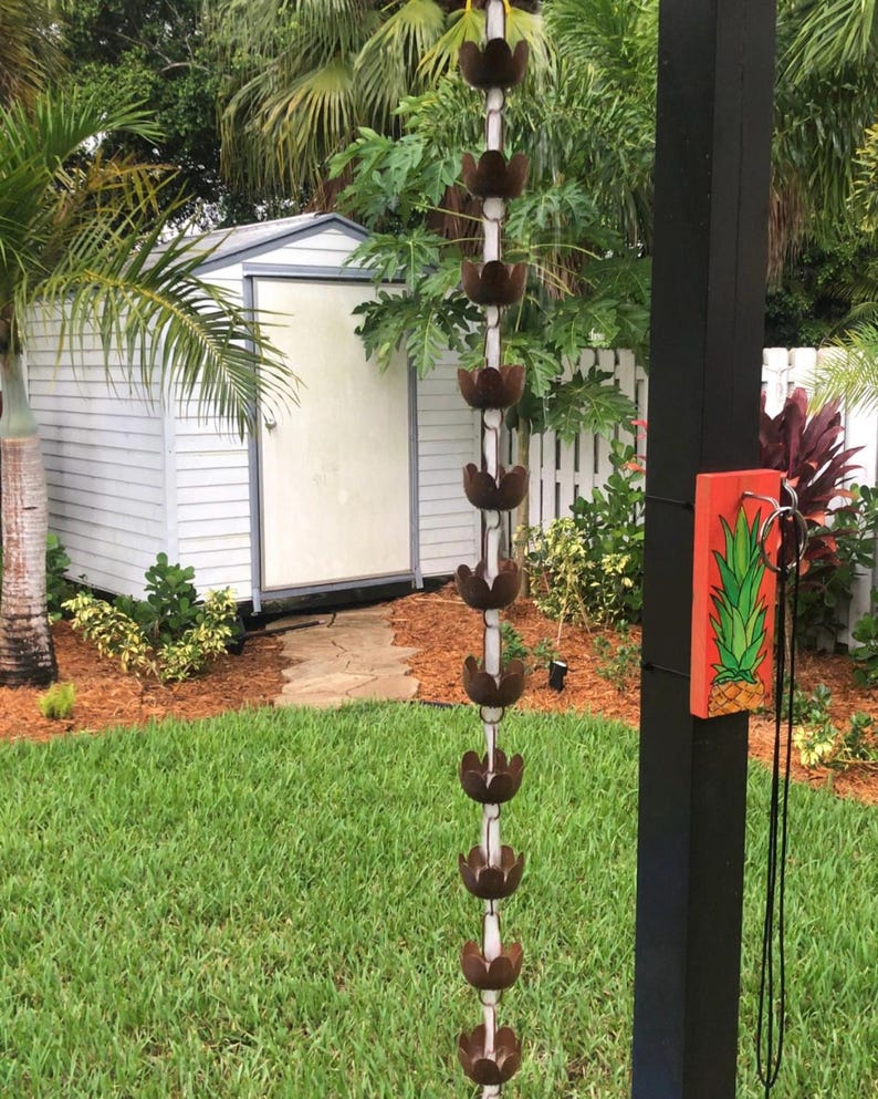 May include: A white shed with a brown door in a garden with a green lawn. A brown metal rain chain hangs from a black post with a red and green pineapple design on a wooden sign.