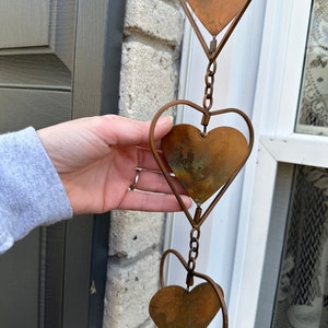 May include: A vertical hanging decoration featuring a series of interconnected metal hearts. Each heart consists of a solid, weathered copper-toned plate framed by a thin, curved wire outline, with small metal chain links connecting each unit.