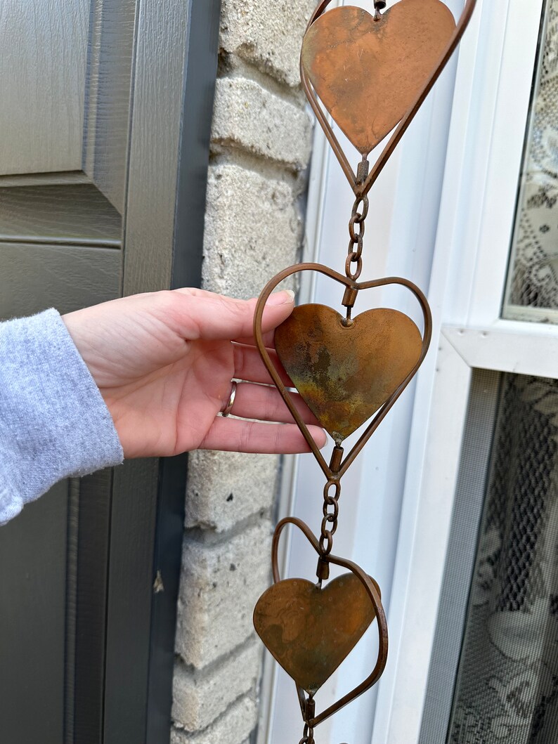 May include: A vertical hanging decoration featuring a series of interconnected metal hearts. Each heart consists of a solid, weathered copper-toned plate framed by a thin, curved wire outline, with small metal chain links connecting each unit.