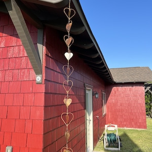 May include: A vertical rain chain hanging from the roof eave, constructed from a series of interconnected copper-colored metal hearts. Each heart frame contains a smaller, solid heart-shaped leaf element suspended within its center, creating a repeating decorative pattern that extends downward along the side of a red building.