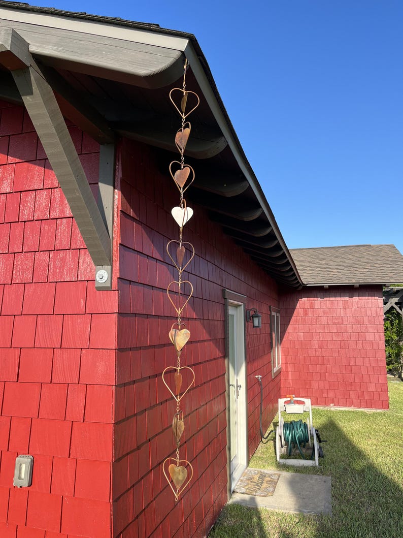 May include: A vertical rain chain hanging from the roof eave, constructed from a series of interconnected copper-colored metal hearts. Each heart frame contains a smaller, solid heart-shaped leaf element suspended within its center, creating a repeating decorative pattern that extends downward along the side of a red building.