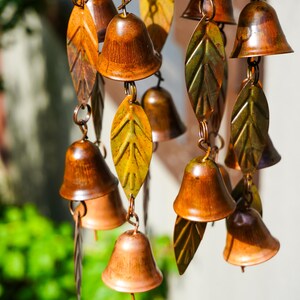 Puede incluir: Un carill&oacute;n de viento con m&uacute;ltiples campanas peque&ntilde;as de metal pulido color cobre, intercaladas con hojas decorativas de metal texturizado en tonos verde y amarillo. Los componentes est&aacute;n unidos por finos anillos met&aacute;licos en cascada.