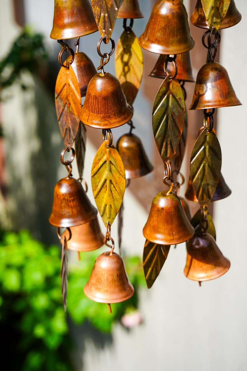 Puede incluir: Un carill&oacute;n de viento con m&uacute;ltiples campanas peque&ntilde;as de metal pulido color cobre, intercaladas con hojas decorativas de metal texturizado en tonos verde y amarillo. Los componentes est&aacute;n unidos por finos anillos met&aacute;licos en cascada.