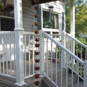 May include: A white porch with a white railing and a copper rain chain hanging from the roof. The rain chain is made of a series of copper cups that are connected by a chain. The rain chain is hanging in front of a window with a white frame.