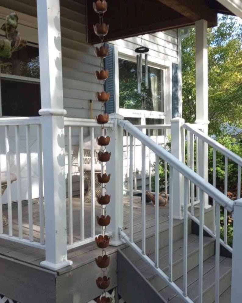 May include: A white porch with a white railing and a copper rain chain hanging from the roof. The rain chain is made of a series of copper cups that are connected by a chain. The rain chain is hanging in front of a window with a white frame.