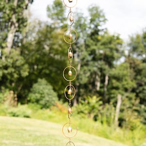 May include: A decorative wind chime featuring a series of small, gold-colored bells suspended from a chain. The bells are attached to gold-colored rings of varying sizes. The background is a blurred view of a green lawn and trees.