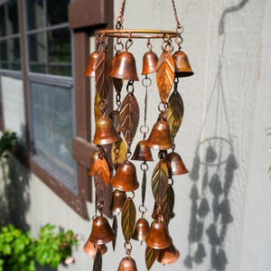 Puede incluir: Un carill&oacute;n de viento colgante con un marco circular de metal en tono cobre suspendido por cadenas. El carill&oacute;n consta de m&uacute;ltiples adornos peque&ntilde;os en forma de campana y piezas met&aacute;licas alargadas en forma de hoja en tonos bronce y cobre.