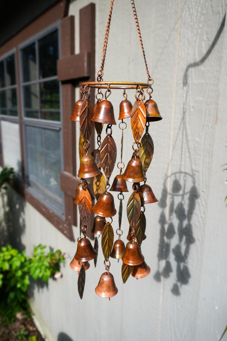 Puede incluir: Un carill&oacute;n de viento colgante con un marco circular de metal en tono cobre suspendido por cadenas. El carill&oacute;n consta de m&uacute;ltiples adornos peque&ntilde;os en forma de campana y piezas met&aacute;licas alargadas en forma de hoja en tonos bronce y cobre.