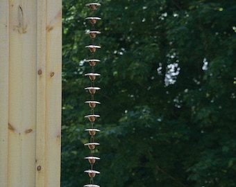 May include: A copper rain chain hangs from the edge of a wooden roof. The chain is made up of a series of connected cups, each with a slightly different shape. The chain is hanging against a backdrop of green trees.