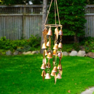 Puede incluir: Un carill&oacute;n de viento de tono cobre con m&uacute;ltiples adornos peque&ntilde;os en forma de campana y piezas de metal con forma de hoja suspendidas de un marco circular mediante cadenas finas, con un gancho en la parte superior para colgar.