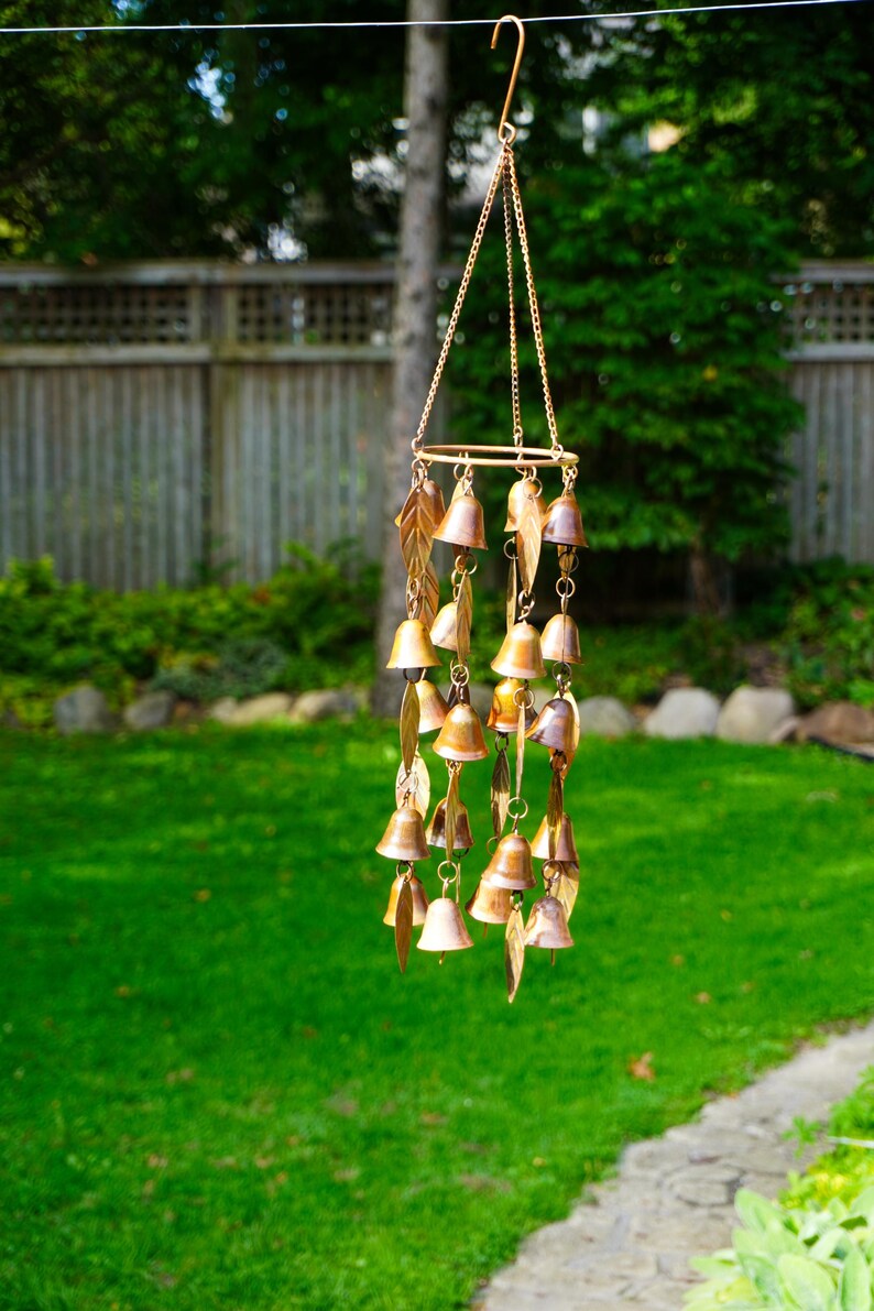 Puede incluir: Un carill&oacute;n de viento de tono cobre con m&uacute;ltiples adornos peque&ntilde;os en forma de campana y piezas de metal con forma de hoja suspendidas de un marco circular mediante cadenas finas, con un gancho en la parte superior para colgar.