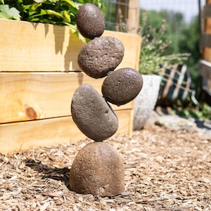 May include: A stack of brown river rocks balanced on top of each other. The rocks are various sizes and shapes, with a smooth, weathered texture. The arrangement is set outdoors on a bed of wood chips, with a wooden planter box and greenery in the background.