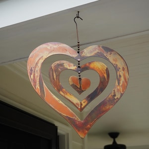 May include: A copper-colored metal heart-shaped wind chime with three concentric hearts. The chime is hanging from a hook on a white ceiling.