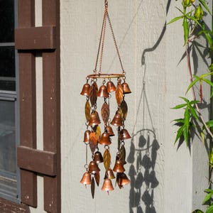 Puede incluir: Un carill&oacute;n de viento de color cobre con m&uacute;ltiples campanas peque&ntilde;as y adornos met&aacute;licos en forma de hoja suspendidos de un marco circular mediante cadenas finas. La pieza decorativa cuelga de un gancho de metal negro y tiene acabado met&aacute;lico.