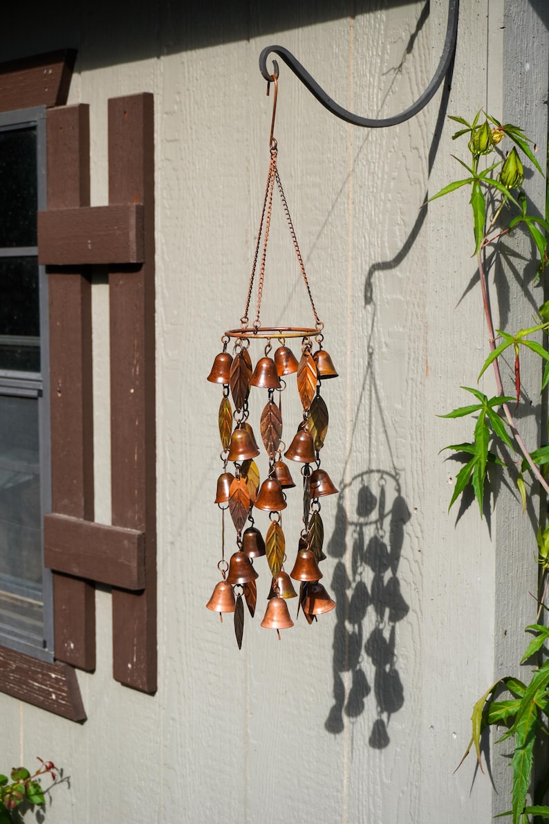 Puede incluir: Un carill&oacute;n de viento de color cobre con m&uacute;ltiples campanas peque&ntilde;as y adornos met&aacute;licos en forma de hoja suspendidos de un marco circular mediante cadenas finas. La pieza decorativa cuelga de un gancho de metal negro y tiene acabado met&aacute;lico.