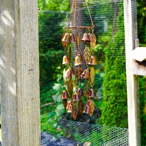Puede incluir: Un carill&oacute;n de viento de tono cobre que presenta m&uacute;ltiples adornos peque&ntilde;os en forma de campana y recortes de metal en forma de hoja suspendidos de una barra horizontal mediante cadenas finas. Los componentes met&aacute;licos tienen un acabado c&aacute;lido y reflectante.
