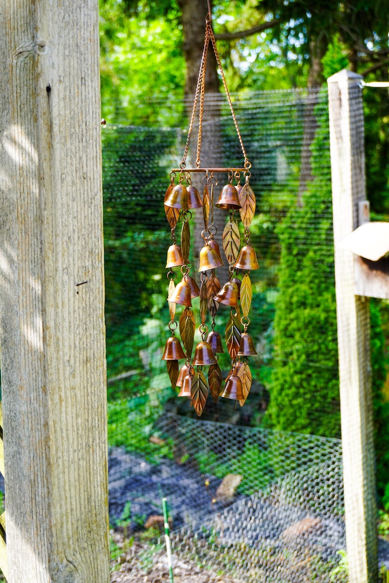 Puede incluir: Un carill&oacute;n de viento de tono cobre que presenta m&uacute;ltiples adornos peque&ntilde;os en forma de campana y recortes de metal en forma de hoja suspendidos de una barra horizontal mediante cadenas finas. Los componentes met&aacute;licos tienen un acabado c&aacute;lido y reflectante.