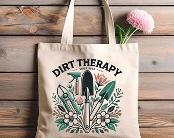 May include: A white canvas tote bag with a green and brown design featuring gardening tools and flowers. The text "Dirt Therapy Since 2018" is printed on the bag.