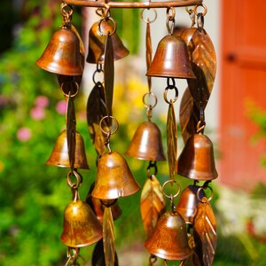 Puede incluir: Un carill&oacute;n de viento de color cobre con m&uacute;ltiples adornos peque&ntilde;os en forma de campana y piezas met&aacute;licas alargadas en forma de hoja, suspendidas de un marco circular mediante finas cadenas. Los componentes tienen un acabado met&aacute;lico desgastado.