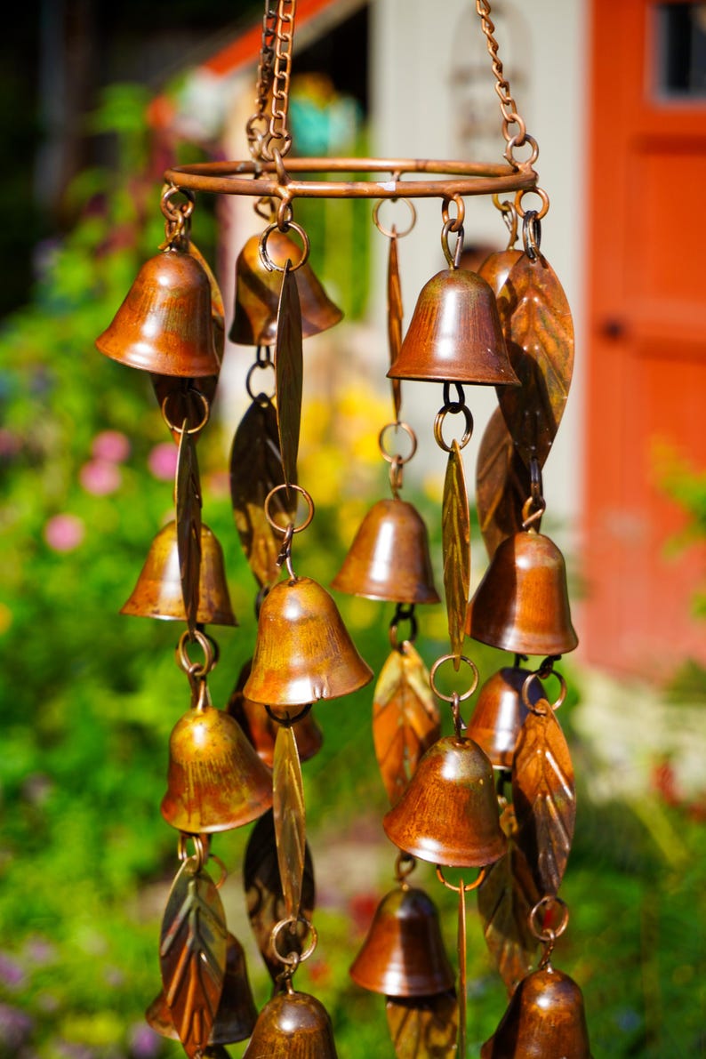 Puede incluir: Un carill&oacute;n de viento de color cobre con m&uacute;ltiples adornos peque&ntilde;os en forma de campana y piezas met&aacute;licas alargadas en forma de hoja, suspendidas de un marco circular mediante finas cadenas. Los componentes tienen un acabado met&aacute;lico desgastado.