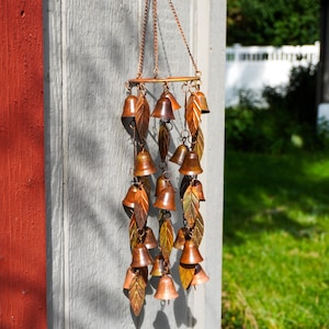 Puede incluir: Un carill&oacute;n de viento colgante que presenta m&uacute;ltiples campanas de metal de color cobre y adornos en forma de hoja suspendidos de un marco circular mediante cadenas finas. El dise&ntilde;o incorpora numerosas piezas peque&ntilde;as en forma de campana.