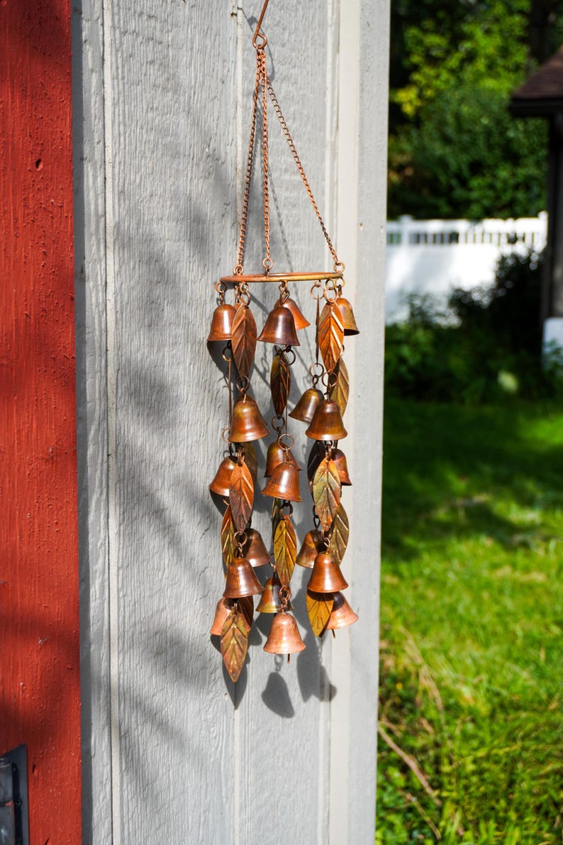 Puede incluir: Un carill&oacute;n de viento colgante que presenta m&uacute;ltiples campanas de metal de color cobre y adornos en forma de hoja suspendidos de un marco circular mediante cadenas finas. El dise&ntilde;o incorpora numerosas piezas peque&ntilde;as en forma de campana.