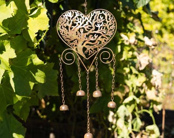 Puede incluir: Un carillón de viento de metal dorado con un diseño en forma de corazón con un motivo de árbol de la vida. El corazón está suspendido de un gancho negro y tiene cadenas colgantes con pequeños adornos en forma de campana. El fondo incluye follaje verde.