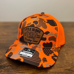 May include: Orange and black camouflage trucker hat with a brown leather patch that says "Little Debbie Hunting Team" and a black and white logo that says "Richardson 112".
