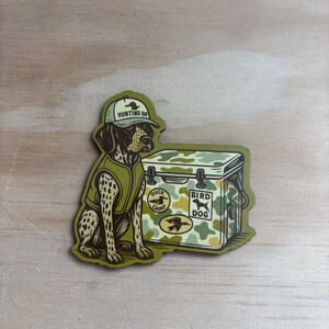 May include: A sticker featuring a hunting dog wearing a cap and vest, sitting next to a camouflage cooler. The cooler has the words "Bird Dog" and "Duck Camp" on it. The dog is brown and white with black spots.
