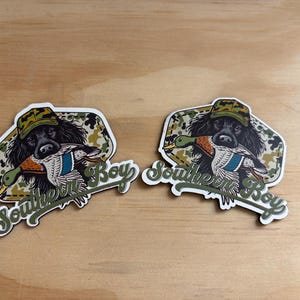 May include: Two stickers featuring a black dog wearing a camouflage hat and holding a duck in its mouth. The stickers have a white border and the words "Southern Boy" in green script.