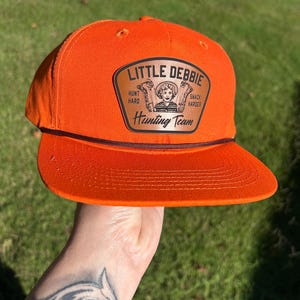 May include: Orange baseball cap with a brown leather patch that says "Little Debbie Hunting Team" with a cartoon image of a woman holding a hunting rifle and the text "Hunt Hard Snack Harder".