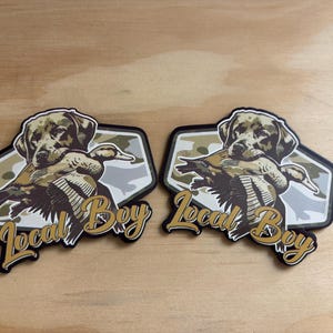May include: Two stickers featuring a Labrador Retriever holding a duck in its mouth. The dog and duck are in shades of brown and tan, set against a camouflage background. The text "Local Boy" is in gold script.